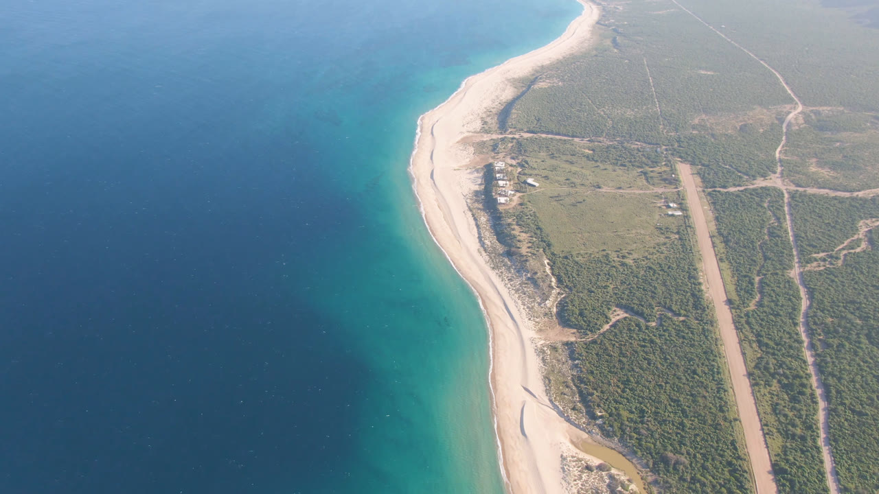 Aerial coastline with blue water, sandy beach, scrubland, and dirt tracks.