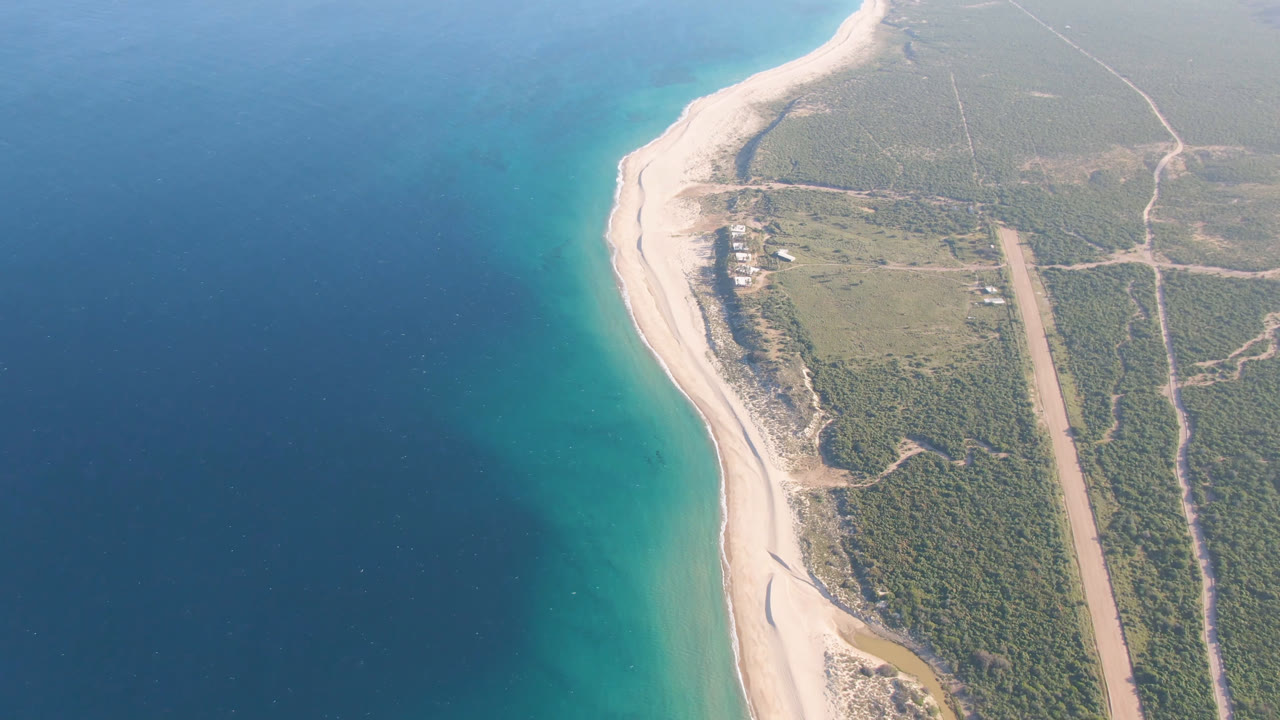 Los Frailes shoreline seen from above, with turquoise shallows along the sand, darker ocean farther out, inland tracks, a long cleared strip, and no visible people.