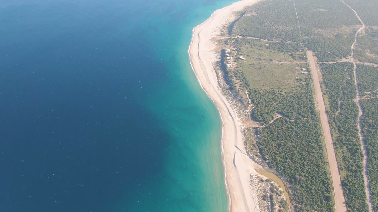 Opening overhead view of the Los Frailes coastline with a pale sand curve, turquoise shallows, dark blue water, green scrubland, dirt tracks, and no visible people.