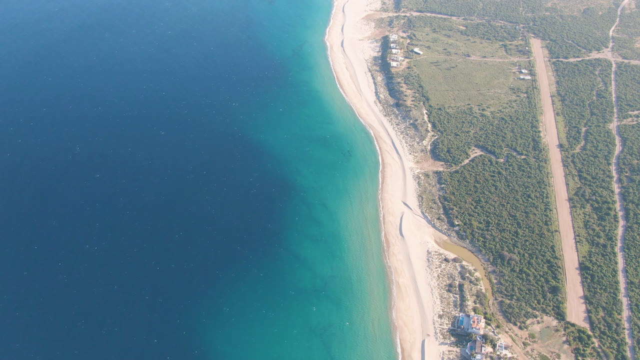 Overhead view of Los Frailes coastline with deep blue water on the left, a pale beach along the center, green scrubland on the right, and no visible people.