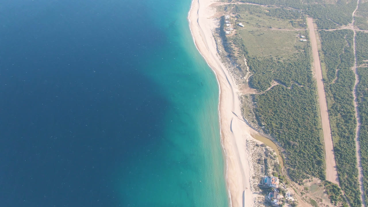 Aerial frame of Los Frailes showing turquoise water near the beach, a narrow sandy shoreline, inland scrub, dirt paths, a straight cleared strip, and no visible people.