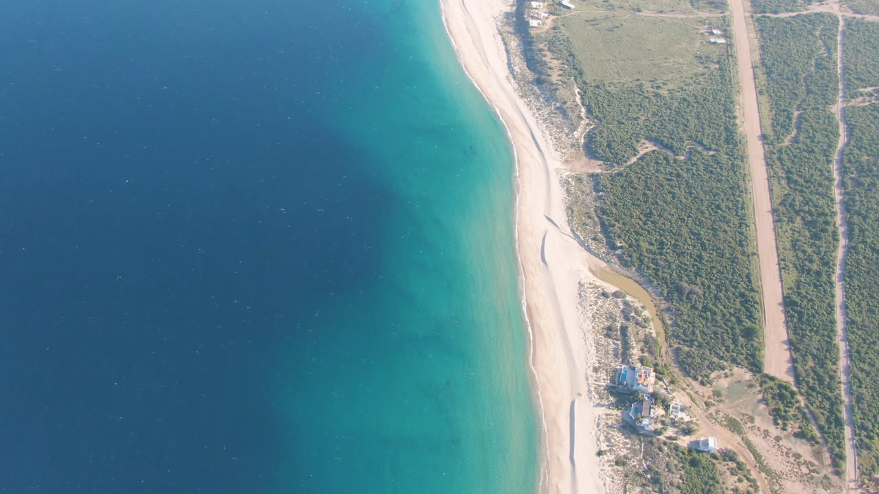 Overhead view of blue water, turquoise shallows, sandy beach, scrubland, road, and small buildings.