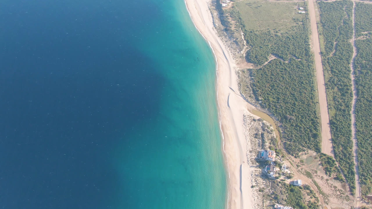 Second aerial frame at Los Frailes shows the beach curving beside turquoise water, with the small building cluster slightly lower in frame and no visible people.