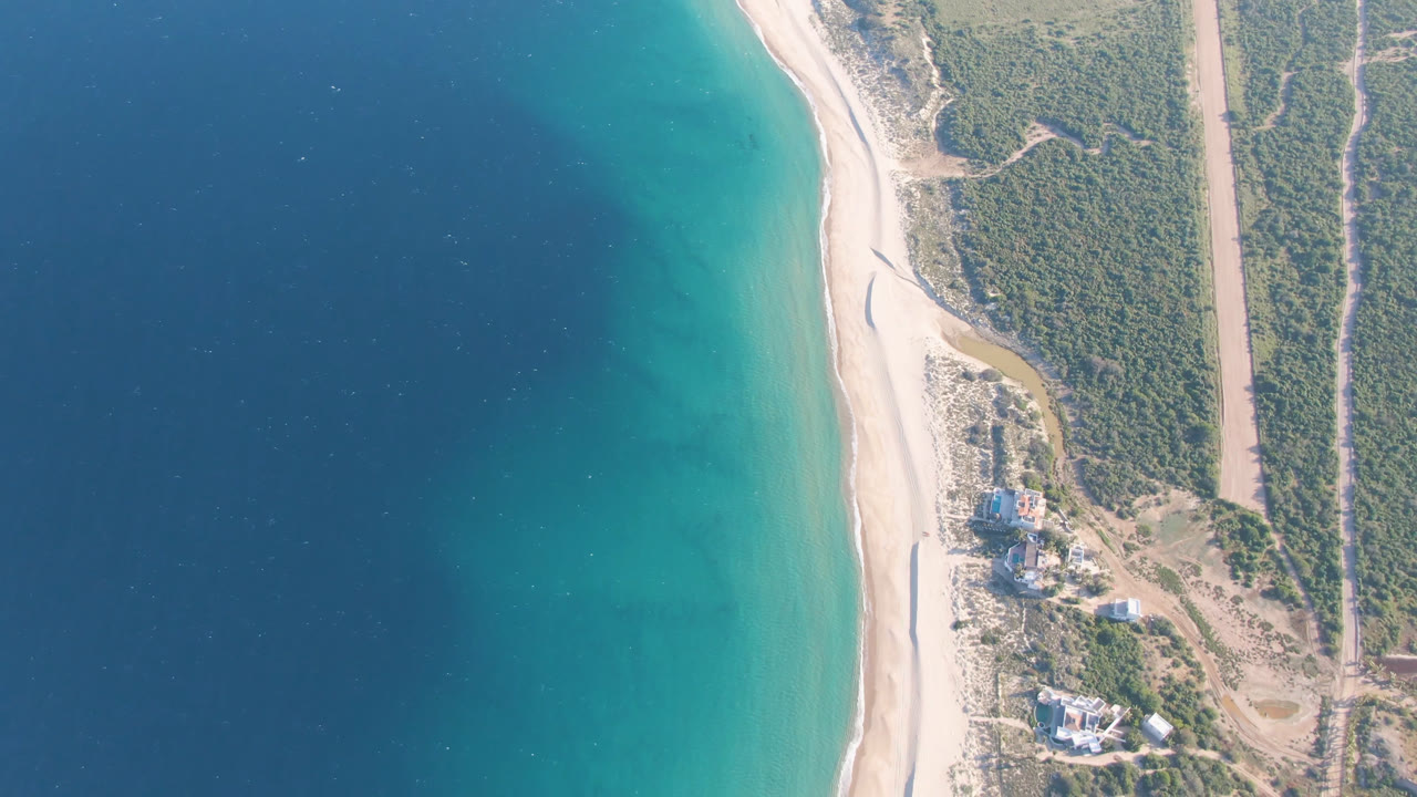 Overhead view of blue ocean, turquoise shallows, pale beach, scrubland, road, and small coastal buildings.
