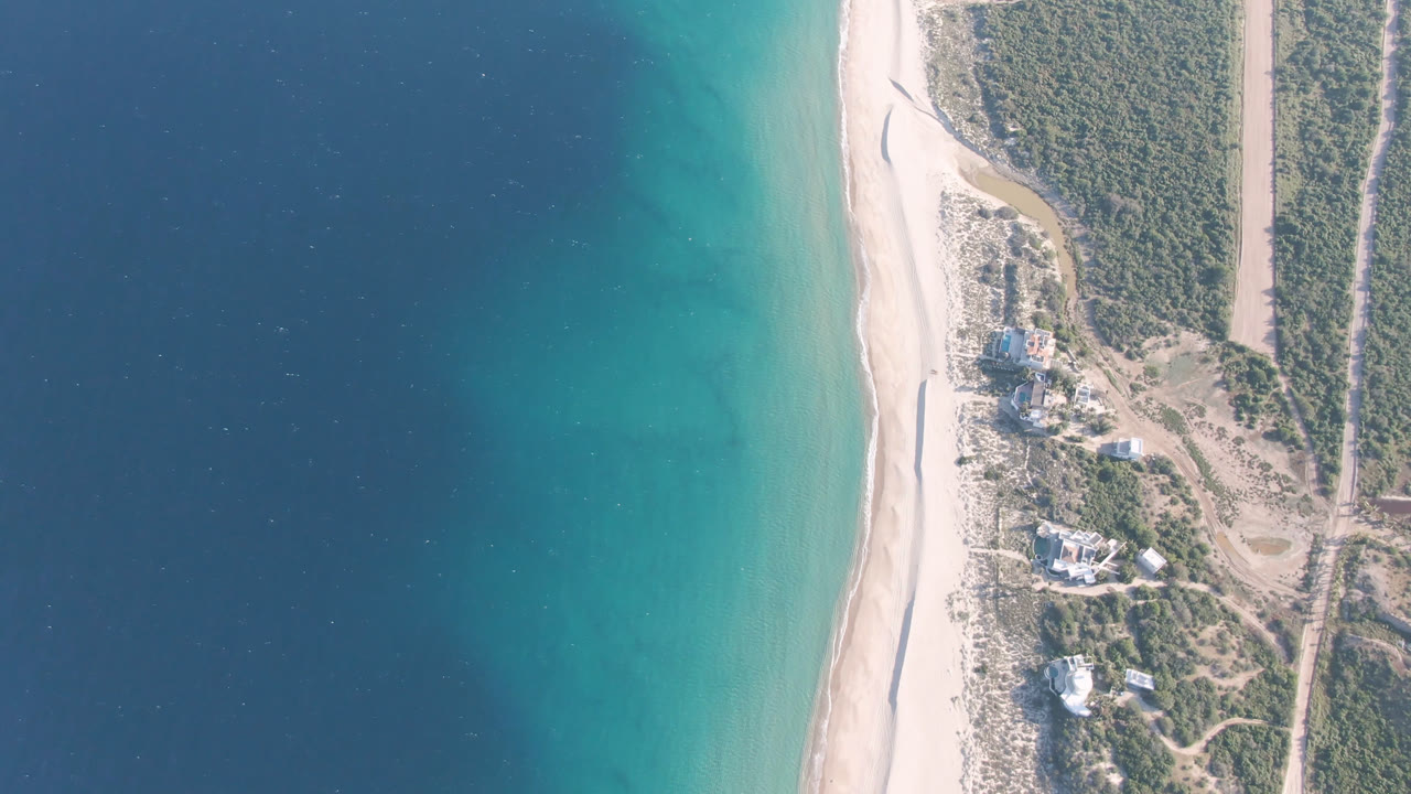 Overhead coastline with blue water, turquoise shallows, beach, scrubland, and scattered buildings.