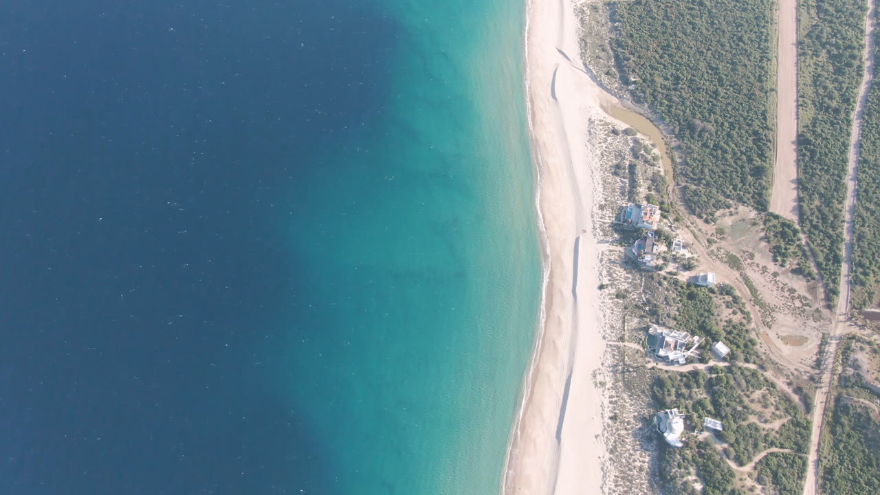 Overhead Los Frailes coastal frame showing a long beach between layered blue-green water and scrub-covered land with dirt roads, with no visible people.