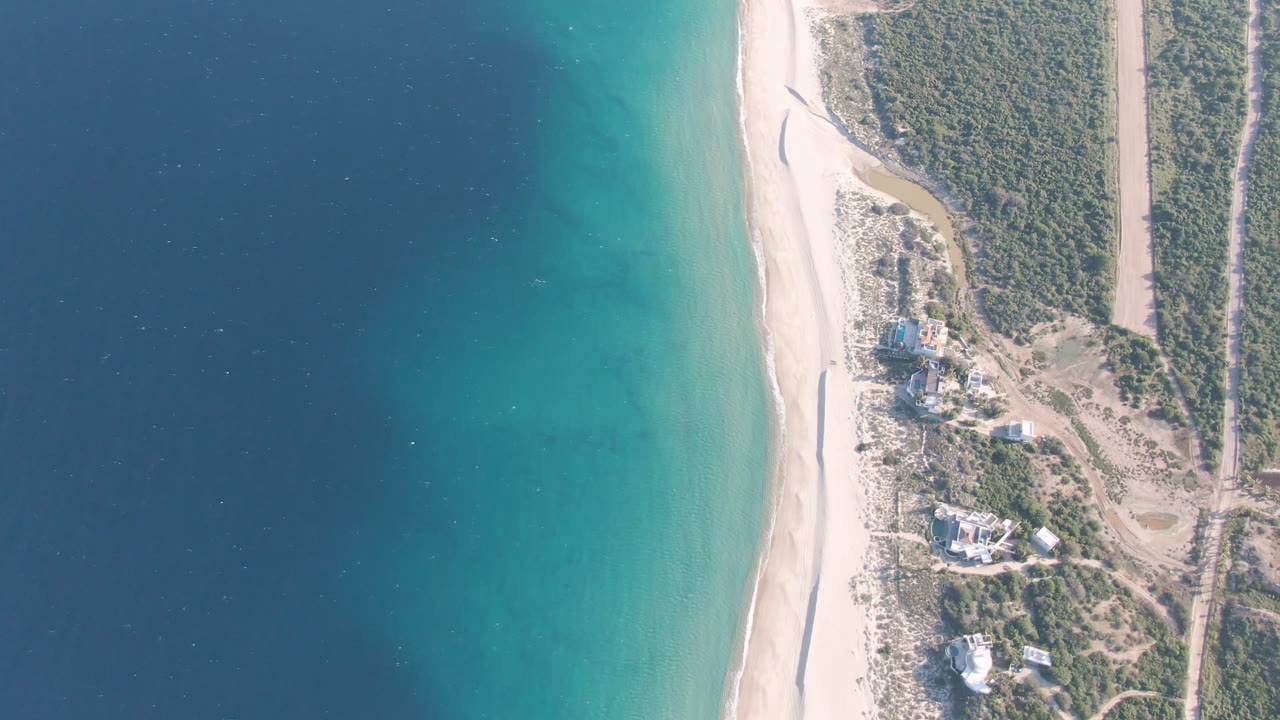 Overhead frame of Los Frailes, Baja California Sur, showing deep blue water fading to turquoise shallows beside a pale beach, coastal scrub, dirt tracks, scattered buildings, and no visible people.