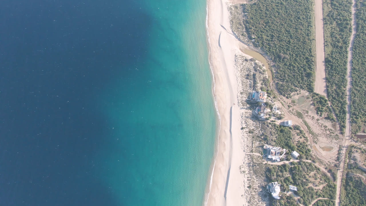 Aerial view over Los Frailes with the shoreline running through the frame, blue-green water on one side, sandy beach, scrubland roads, small structures, and no visible people.
