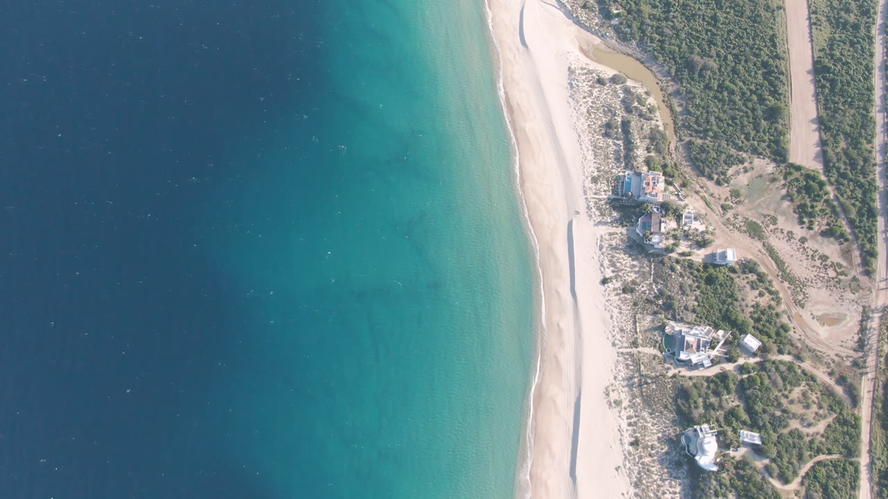 Aerial view of blue water, pale beach, scrub, road, and shoreline buildings.