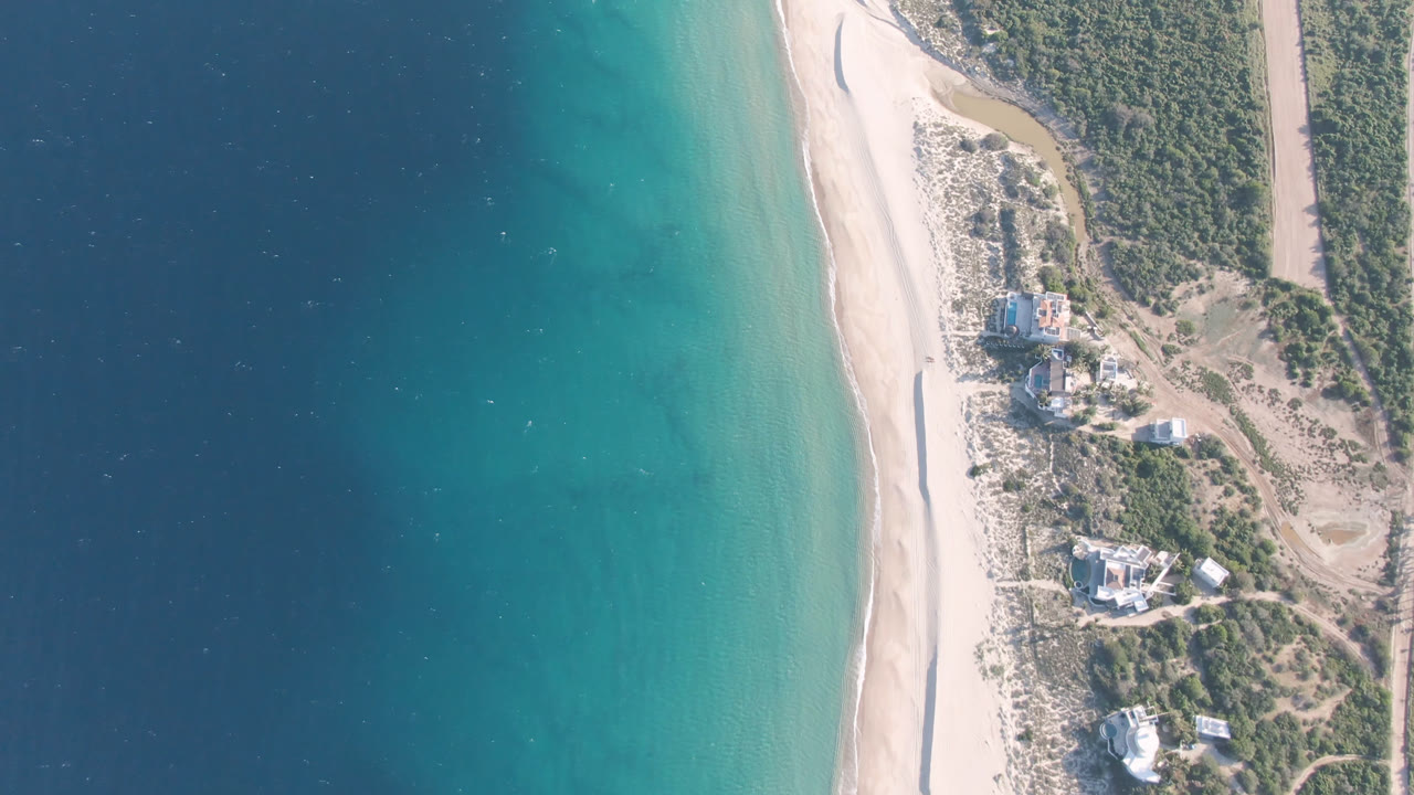 Aerial view at 0:01 over Los Frailes showing the shoreline shifted slightly in frame, with turquoise water beside pale sand, scrub vegetation, an inland road, and no visible people.