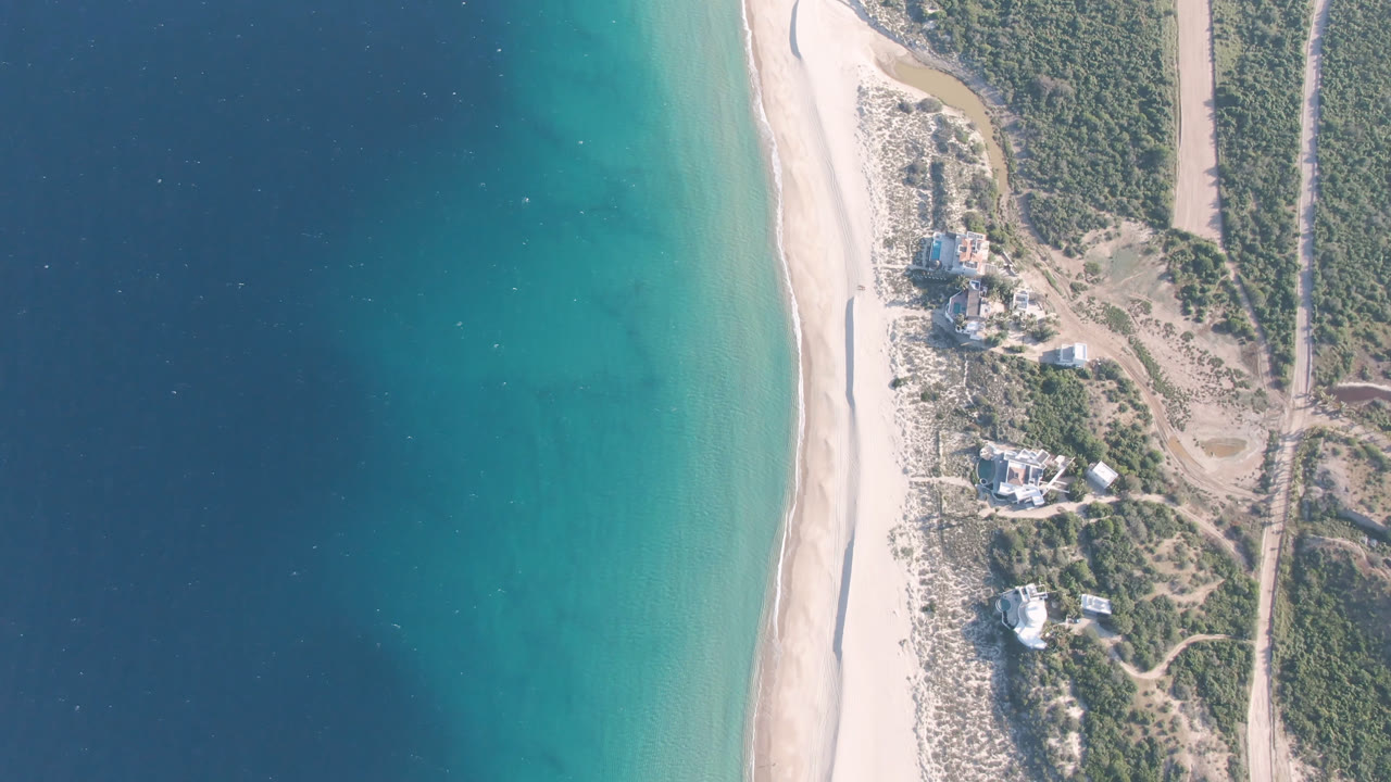 Aerial frame over Los Frailes, Baja California Sur, showing deep blue water shifting to turquoise along a pale beach with scrubland and small buildings inland, no visible people.