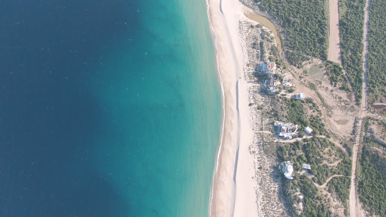 Overhead view of Los Frailes coastline with a wide band of blue-green water, pale sand, dune scrub, a dirt track, and scattered shoreline buildings, no visible people.