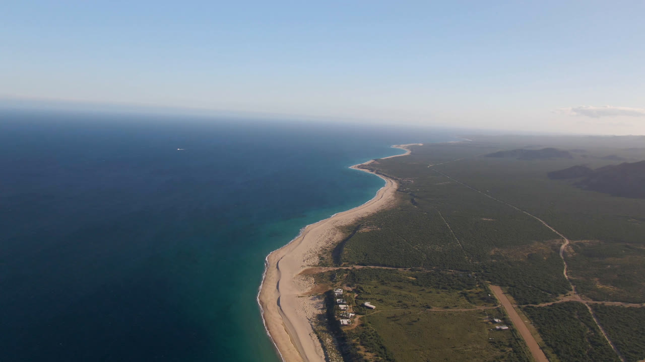 High aerial frame over Los Frailes, Baja California Sur, with deep blue ocean on the left, a pale beach curving into the distance, green scrubland on the right, and no visible people.