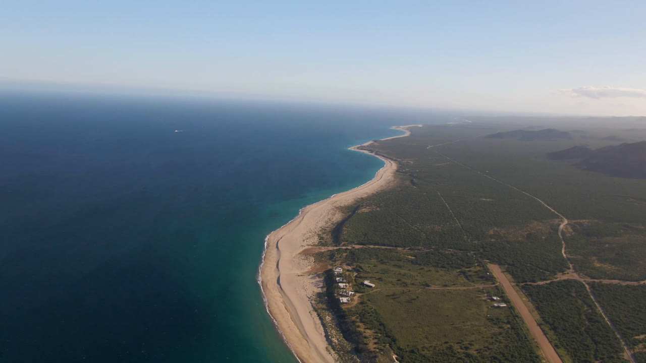 Aerial shoreline view with blue ocean, bright beach, green terrain, roads, and distant hills.