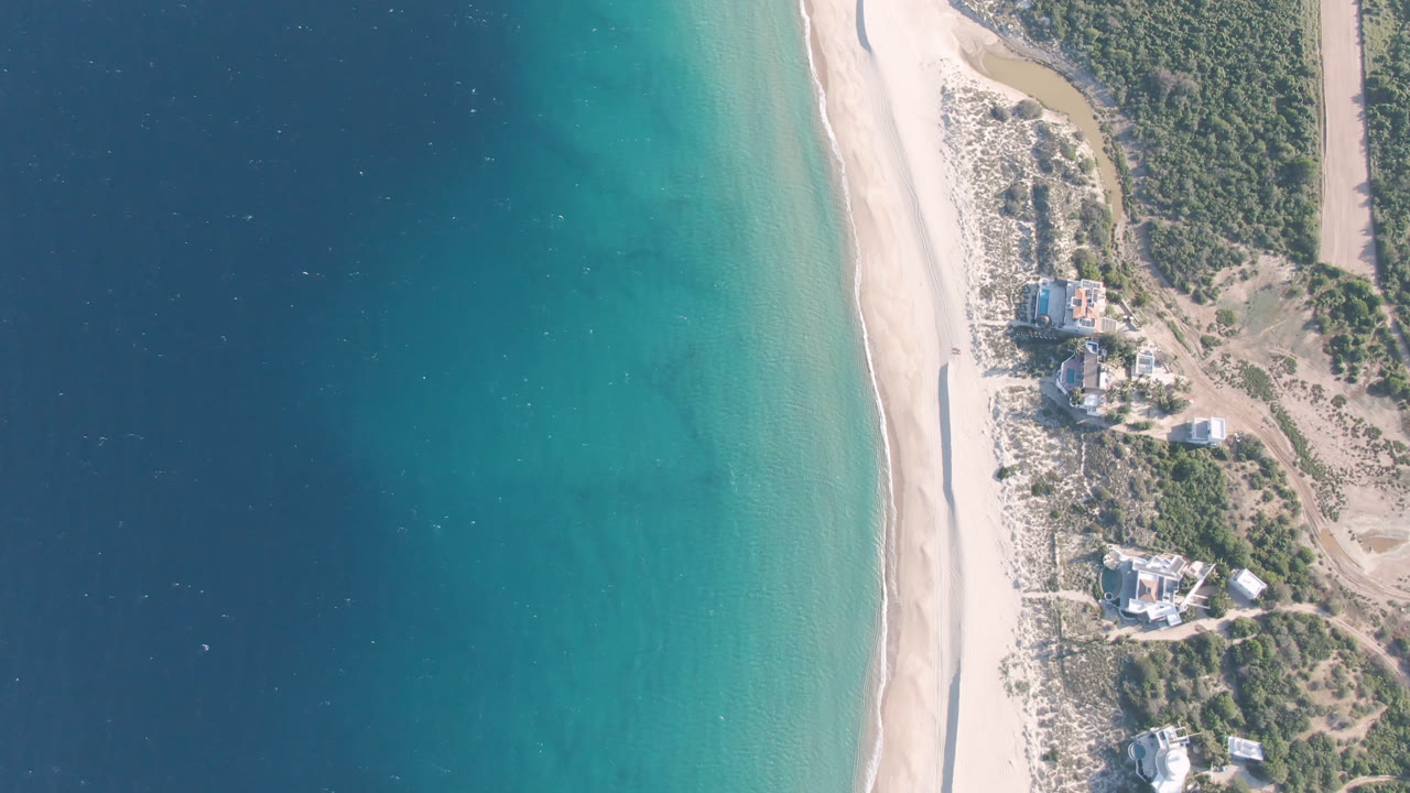 Overhead view of turquoise water, sandy beach, scrub, and coastal buildings.