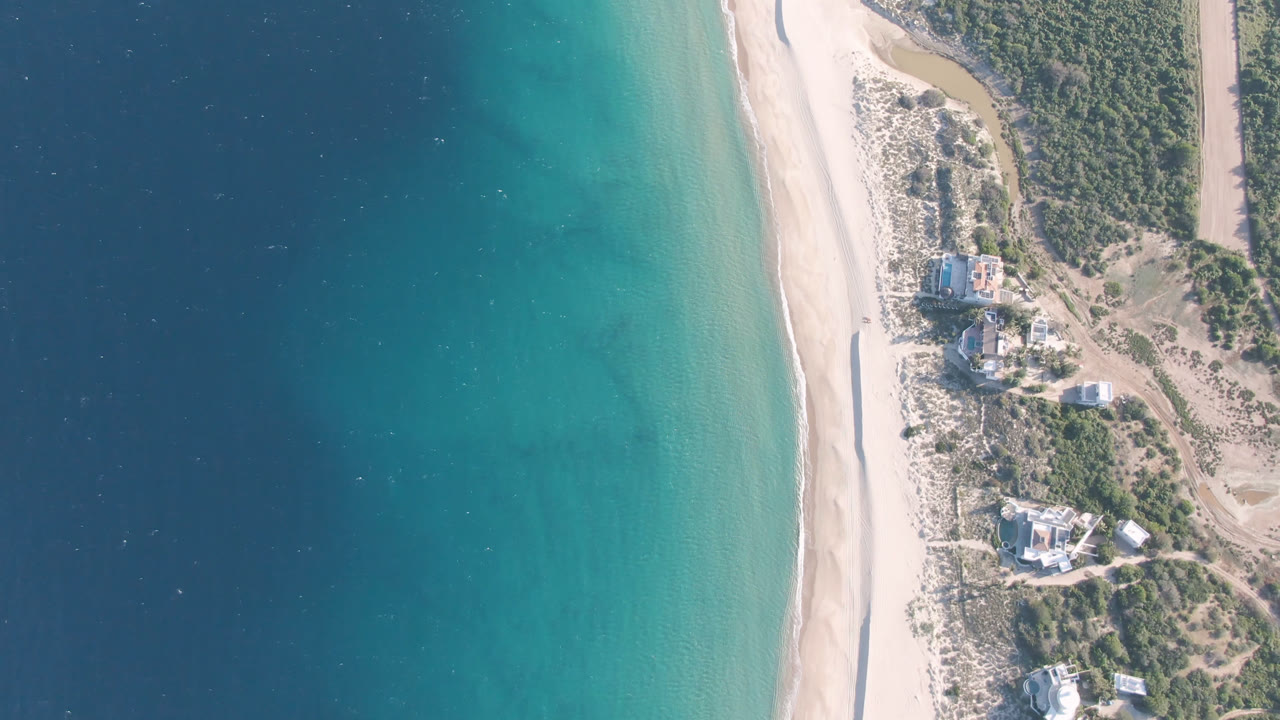Aerial view over Los Frailes with a broad band of blue-green water, a narrow pale beach, inland scrub, dirt paths, and small buildings near the shore, with no visible people.