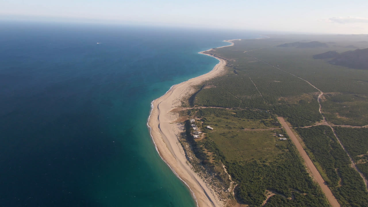 Aerial view of curved beach, blue ocean, scrubland, roads, and small buildings