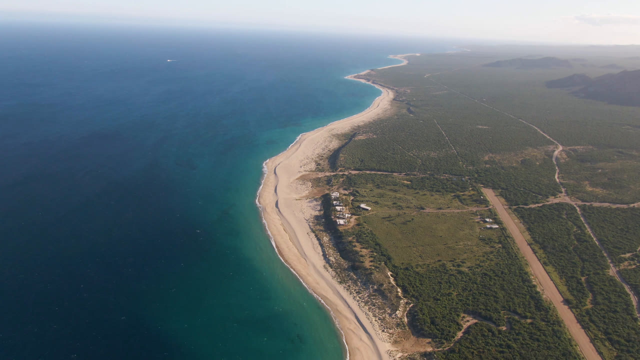 Aerial view of Los Frailes with the sandy shoreline running diagonally through frame, darker ocean offshore, turquoise shallows near the beach, scrubland roads inland, and no visible people.