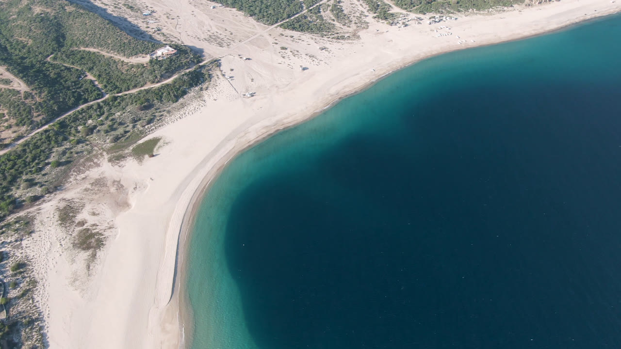 High aerial frame over Los Frailes, Baja California Sur, showing a curved pale beach, scrub dunes, hillside tracks, and dark blue water with no visible people.