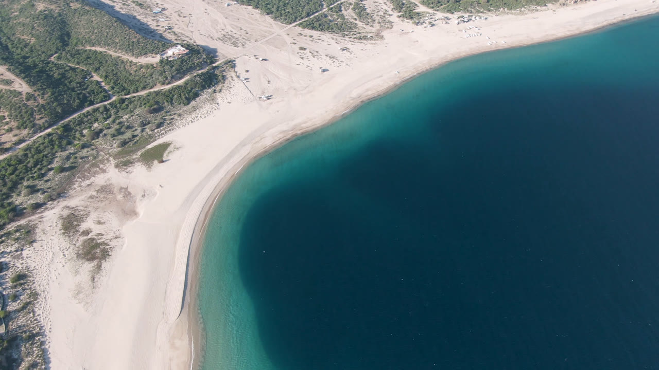 Overhead view of Los Frailes beach with the shoreline sweeping across the frame, green scrubland above the sand, and deep water below with no visible people.