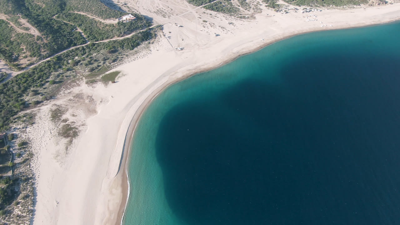 High aerial frame over Los Frailes showing a pale curved beach, turquoise shallows, dark blue water, scrubby dunes, dirt tracks, and no visible people.