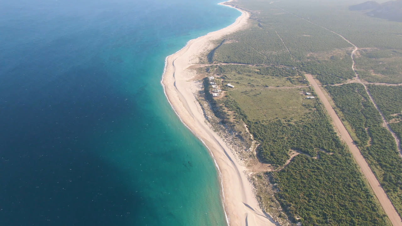 Aerial view of turquoise ocean, sandy beach, scrubland, and dirt road.