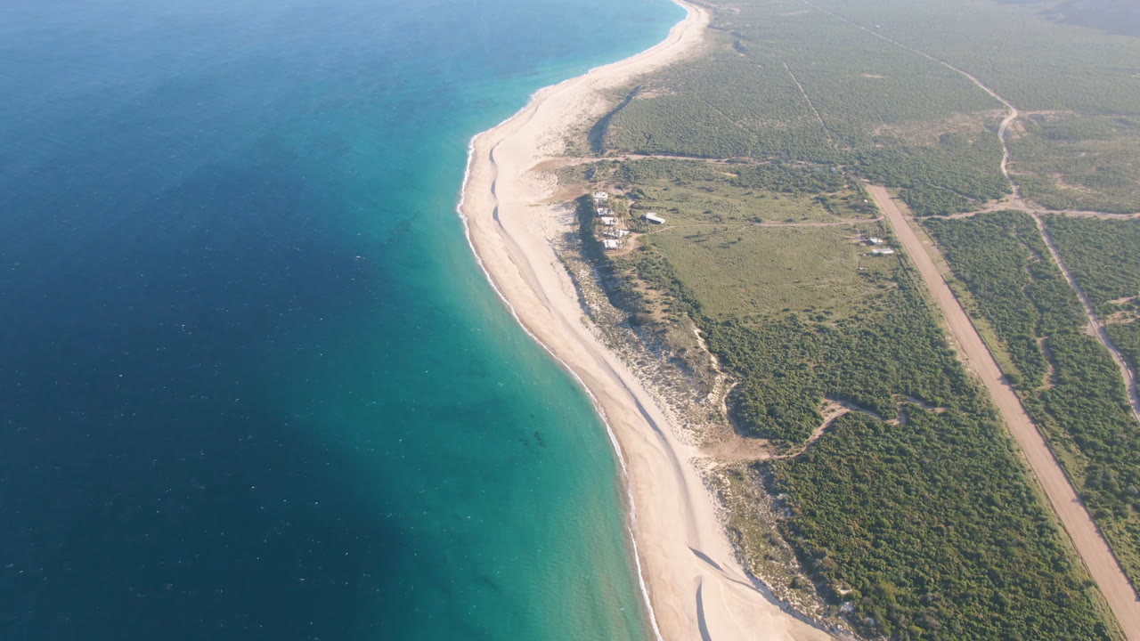 Aerial frame over Los Frailes showing the beach curve more centered, with blue-green shallows along the sand, darker ocean offshore, scrubland inland, and no visible people.