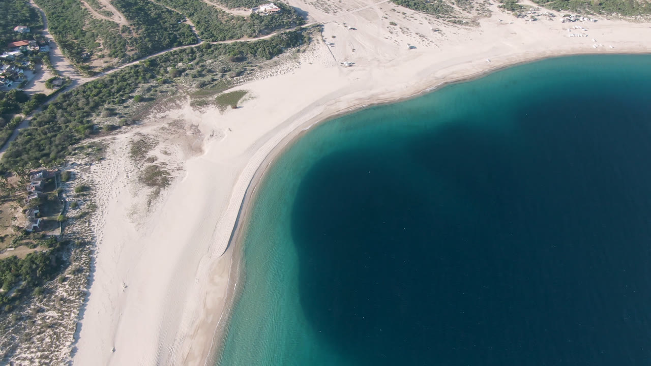 Aerial frame over Los Frailes, Baja California Sur, showing a curved pale beach, turquoise shallows, darker offshore water, scrub dunes, a road, and no visible people.