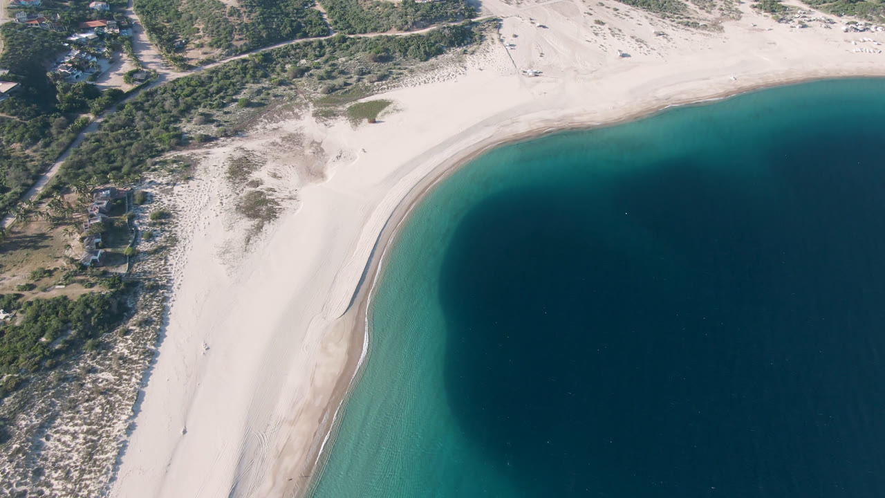 Overhead drone frame of Los Frailes beach with pale sand, turquoise shallows, darker water to the right, scrubby dunes inland, and no visible people.