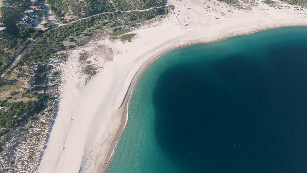 Los Frailes aerial frame with the shoreline running diagonally, a broad pale beach, green-blue nearshore water, darker offshore water, and no visible people.