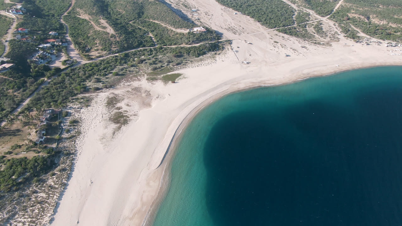 Aerial view at Los Frailes with the shoreline shifted slightly across frame, showing pale sand, a turquoise shallows band, darker bay water, hillside tracks, small buildings, and no visible people.