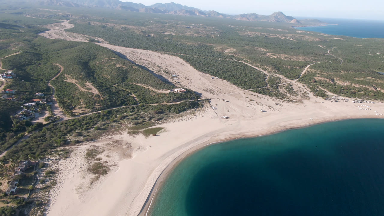Opening aerial frame over Los Frailes, Baja California Sur, with a pale beach curving around dark turquoise water, dry inland wash, green hills, distant mountains, and no visible people.