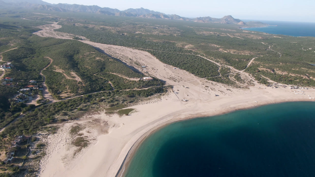 Aerial frame over Los Frailes showing the shoreline slightly farther along the curve, with dark blue water in the foreground, pale sand, inland tracks, low hills, mountains, and no visible people.
