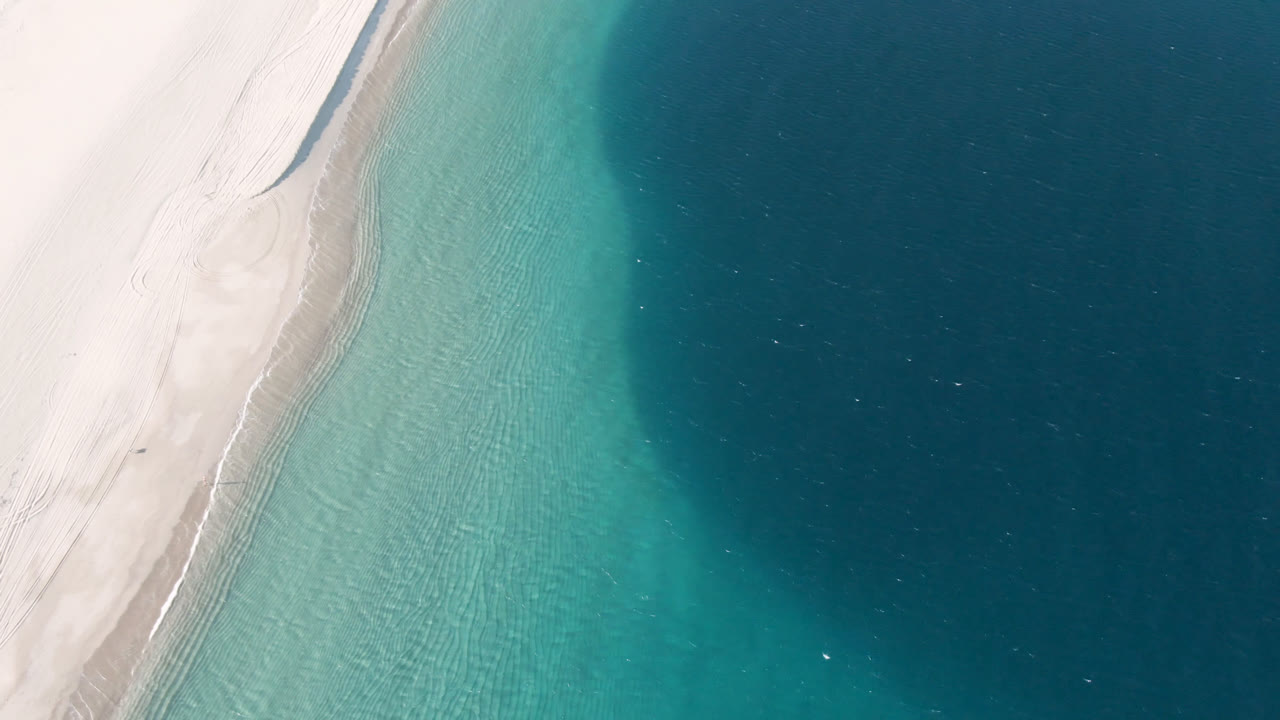 Top-down aerial frame over Los Frailes, Mexico, with pale beach on the left, shallow turquoise water through the center, darker blue water to the right, and no visible people.