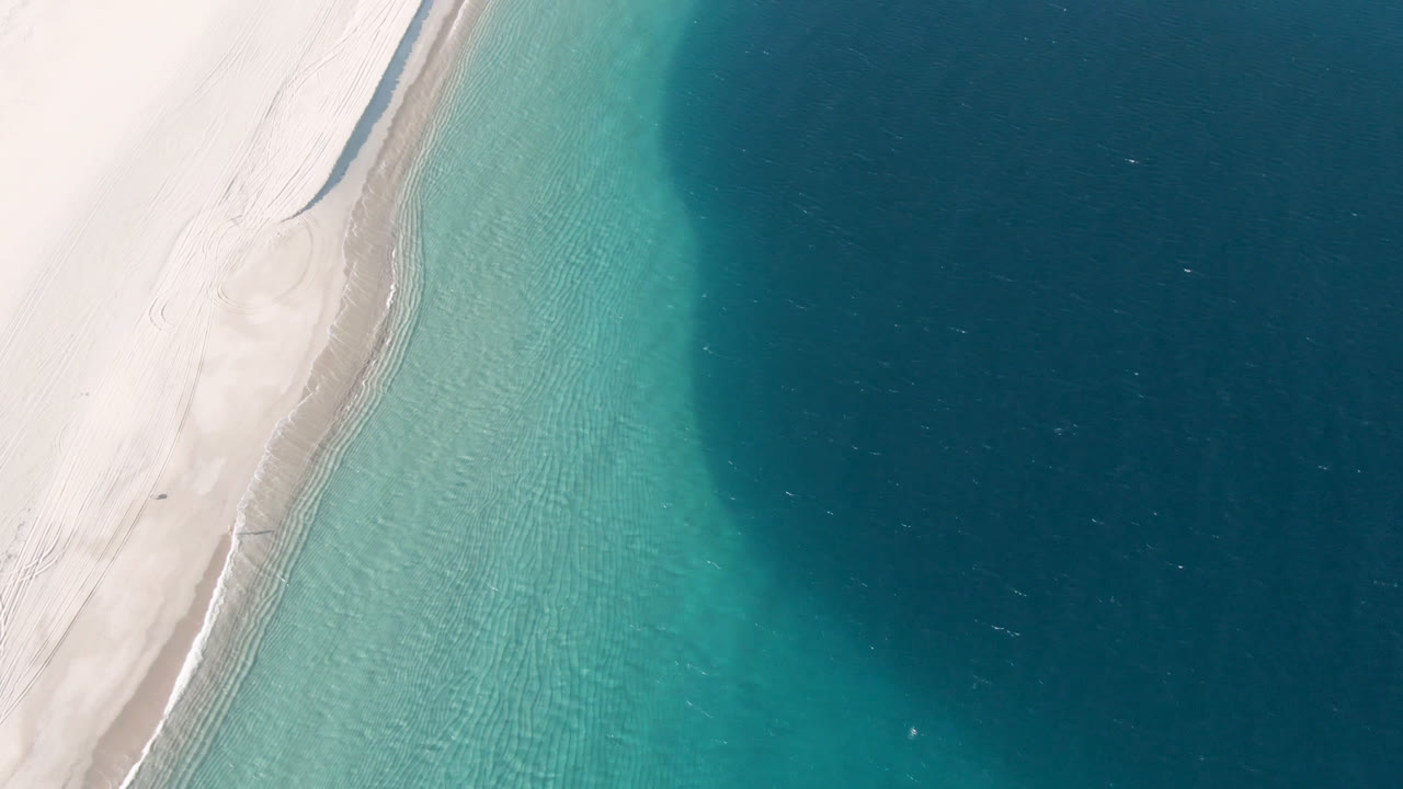 High aerial view at Los Frailes, Mexico, where the sand edge curves beside clear turquoise shallows and the deeper blue water occupies the right side, with no visible people.