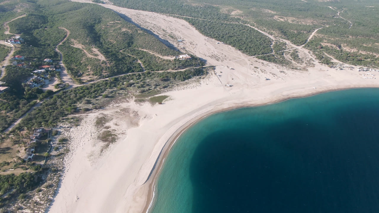 Aerial view of a curved sandy beach, blue bay, and scrub hills.