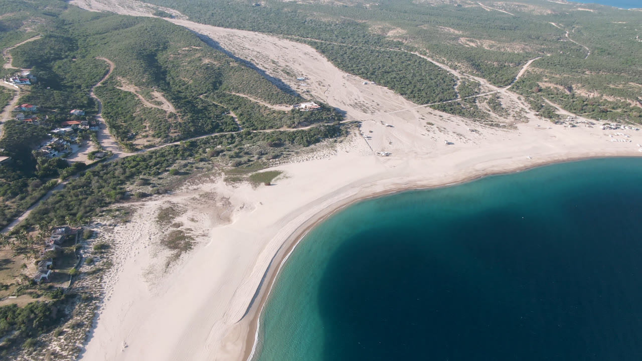Aerial frame over Los Frailes with the beach arc lower in frame, a pale dry wash running inland, scrub hills crossed by dirt tracks, and no visible people.