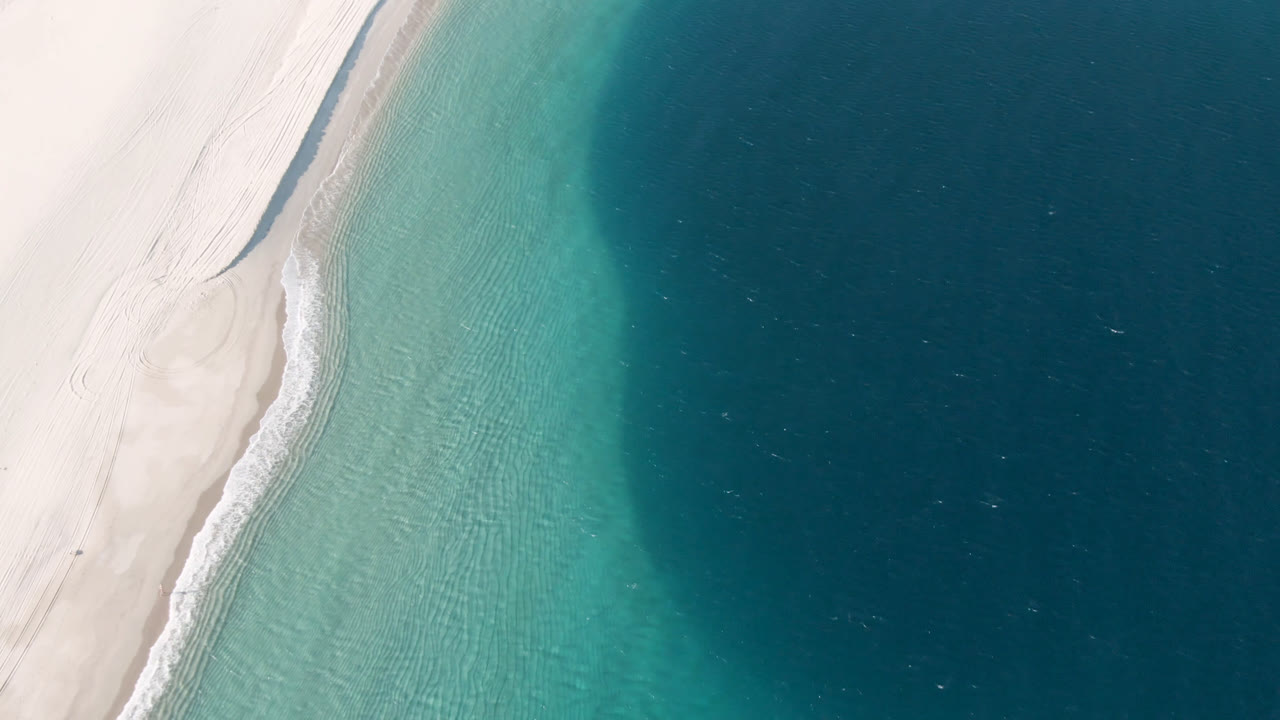 Overhead aerial frame of Los Frailes beach in Baja California Sur, with pale sand on the left, a narrow surf line, turquoise shallows, darker water to the right, and no visible people.