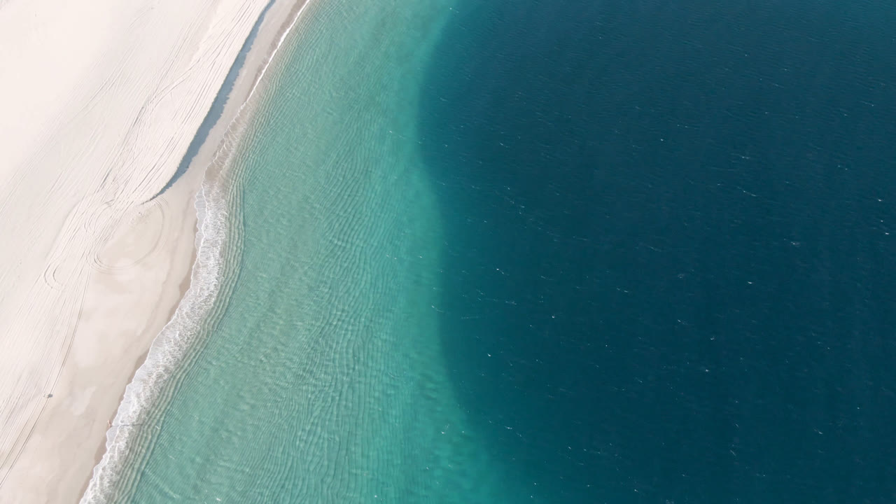 Aerial view over Los Frailes showing the beach edge slightly farther along the curve, with rippled turquoise water, a thin white wave line, deep blue water, and no visible people.