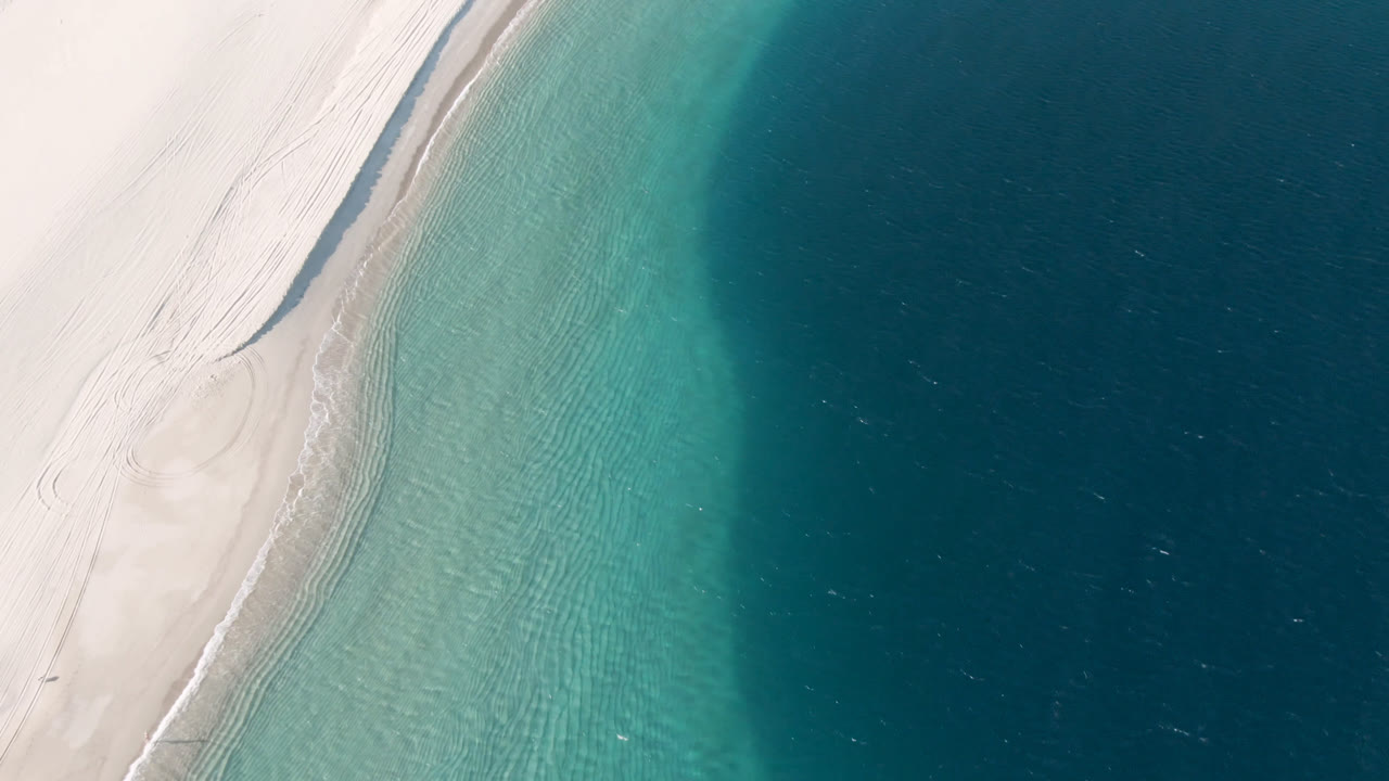 Overhead view at Los Frailes, Baja California Sur, with pale sand curving along the left edge, turquoise shallows in the center, deep blue ocean to the right, and no visible people.
