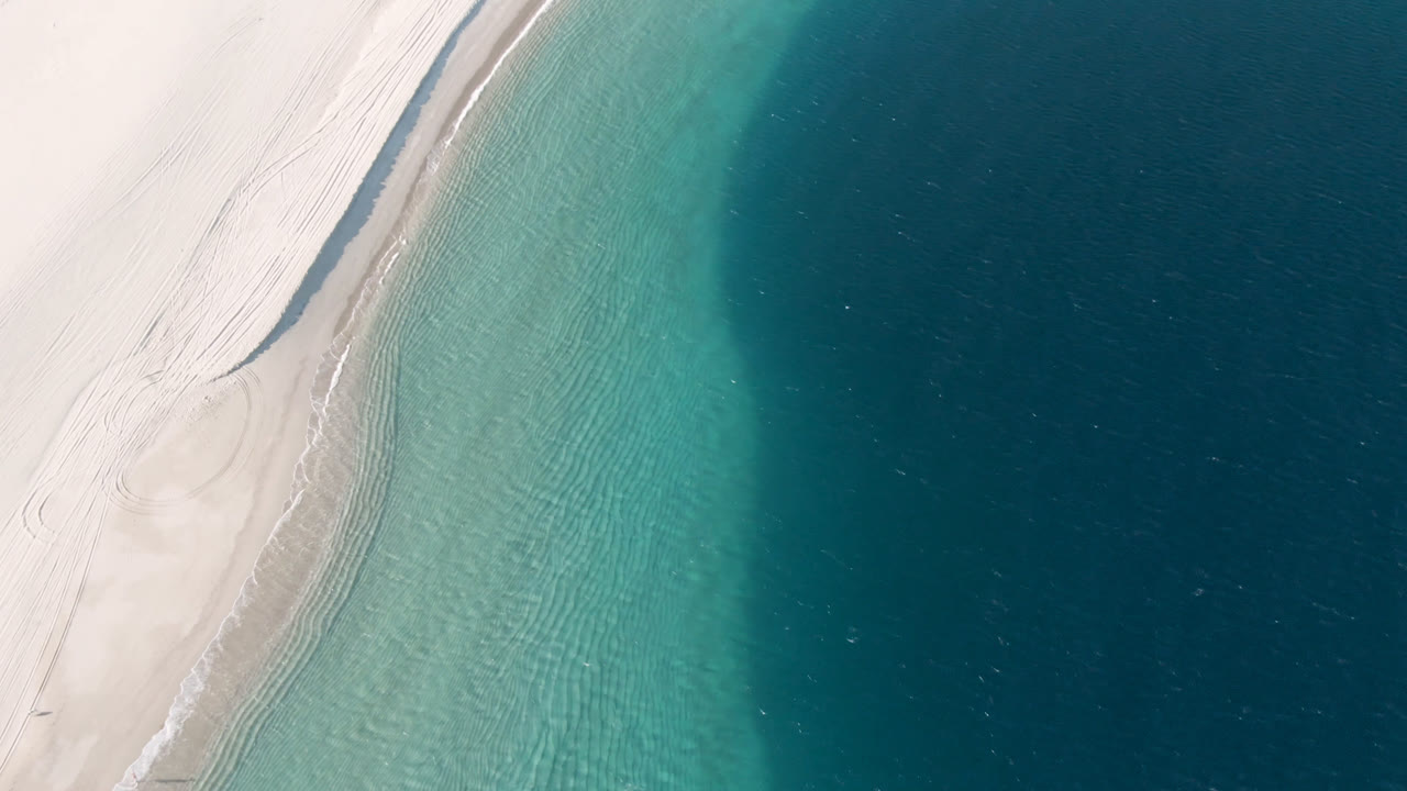 A second overhead frame of Los Frailes shows the sandy beach running diagonally beside rippled turquoise water and a broad band of darker ocean, with no visible people.