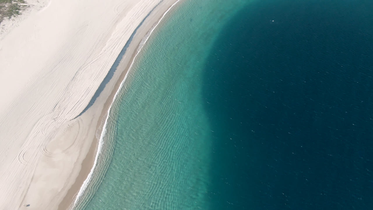 Overhead aerial frame of Los Frailes beach with pale sand at left, a narrow surf line, turquoise shallows, dark blue water to the right, and no visible people.