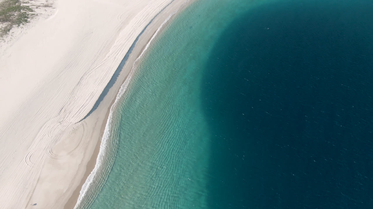Aerial view over Los Frailes shoreline where faint tire or foot tracks run along pale sand, clear green-blue shallows border the beach, and no visible people appear.