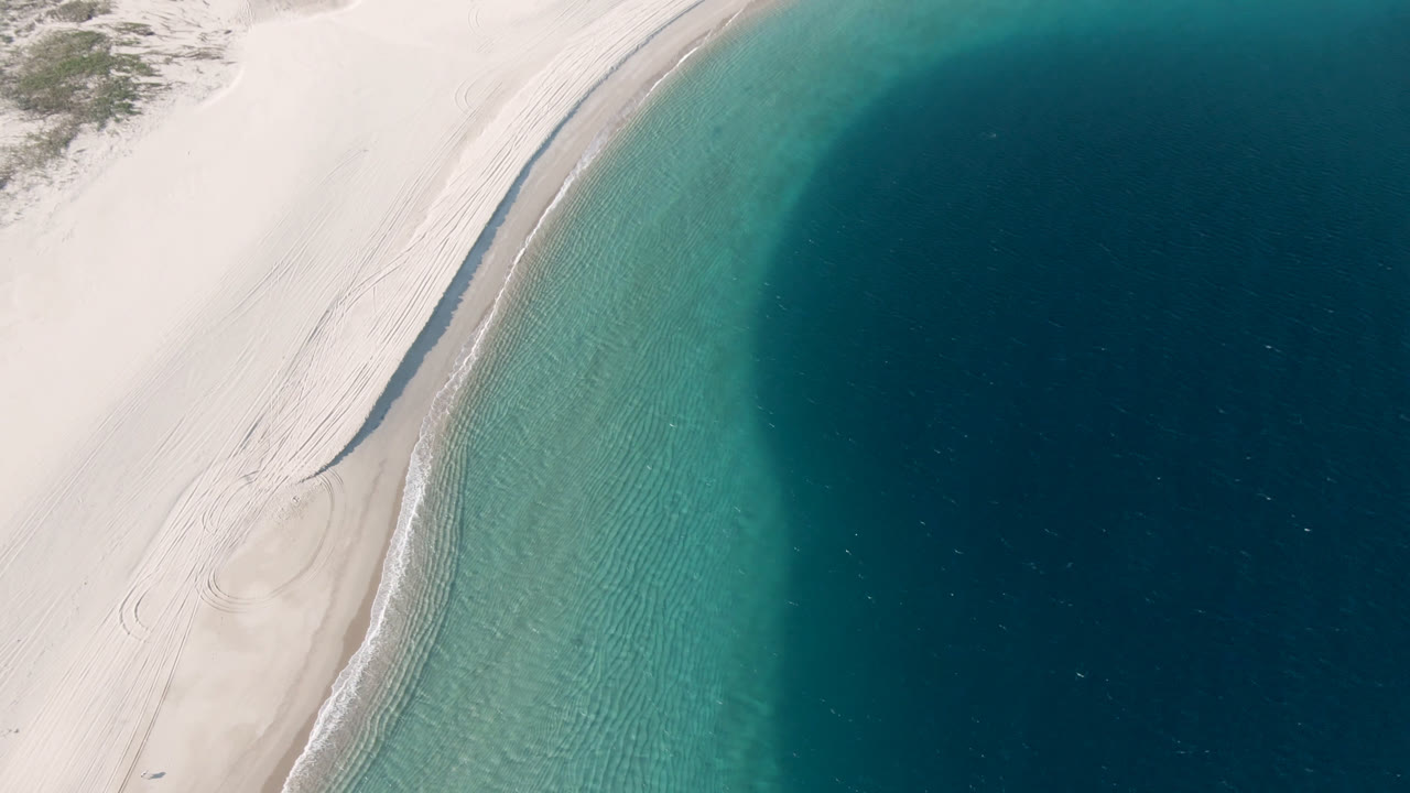 Curved sandy shoreline and rippled turquoise shallows seen from above.