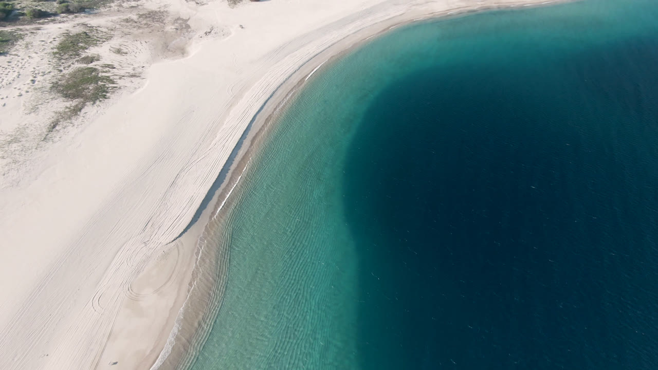 Aerial frame over Los Frailes, Baja California Sur, with pale sand on the left, a curved turquoise shoreline, dark blue water on the right, and no visible people.