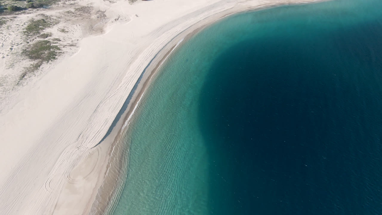A second aerial moment over Los Frailes shows the shoreline slightly farther along the curve, with dune sand, thin surf lines, turquoise shallows, dark water, and no visible people.