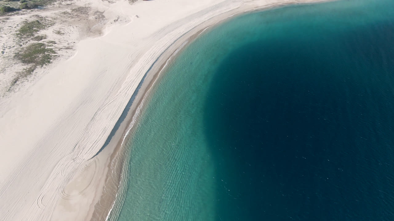 Aerial coastline with pale sand, teal shallows, and deep blue water.