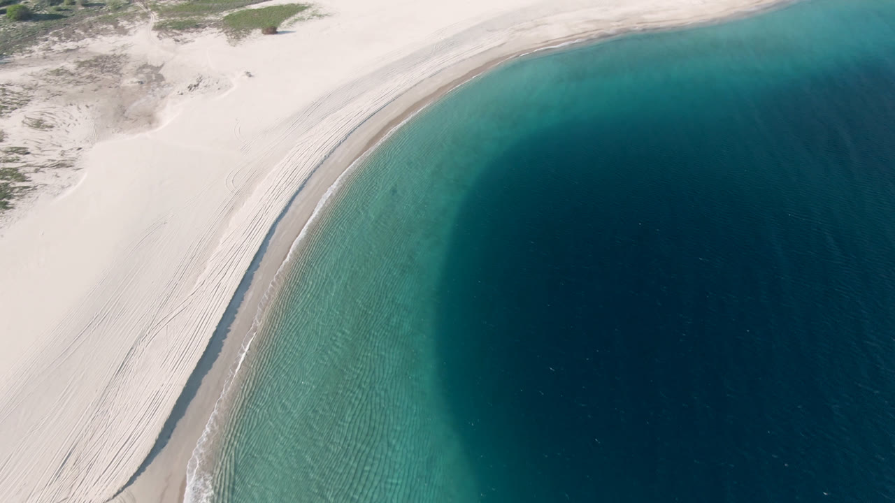 High aerial view over Los Frailes, Baja California Sur, with pale sand curving beside turquoise shallows and darker blue water, no visible people.
