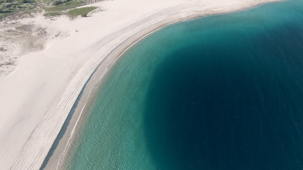 Aerial frame over Los Frailes showing the shoreline running diagonally, with pale sand at left, clear green-blue shallows near shore, and deep blue water to the right, no visible people.