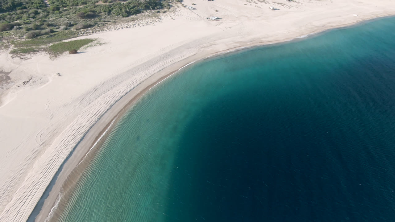 Aerial frame over Los Frailes, Baja California Sur, showing a pale curved beach, shallow green-blue water, darker ocean beyond, dune vegetation, and no visible people.